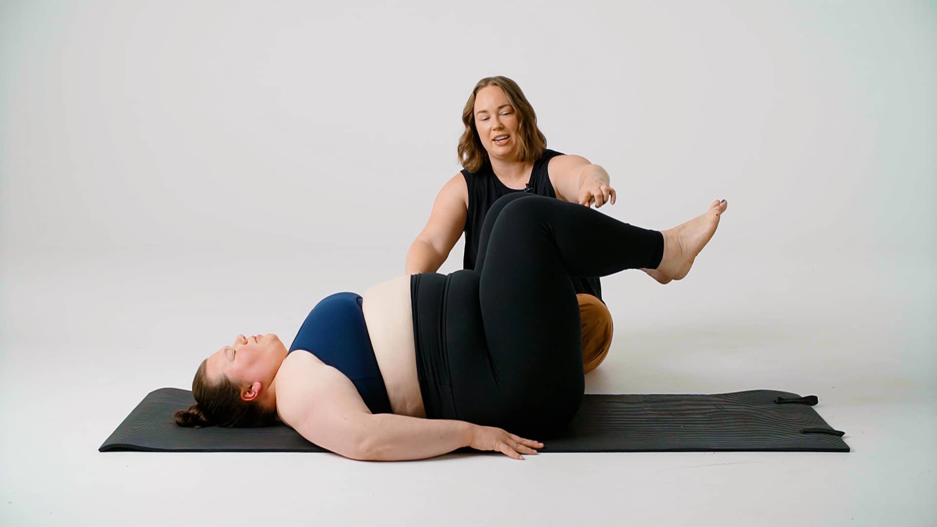Jessie is seated on the ground, performing a diastasis recti assessment, to check the healing on a woman who is laying on her back with her legs in "tabletop" position.