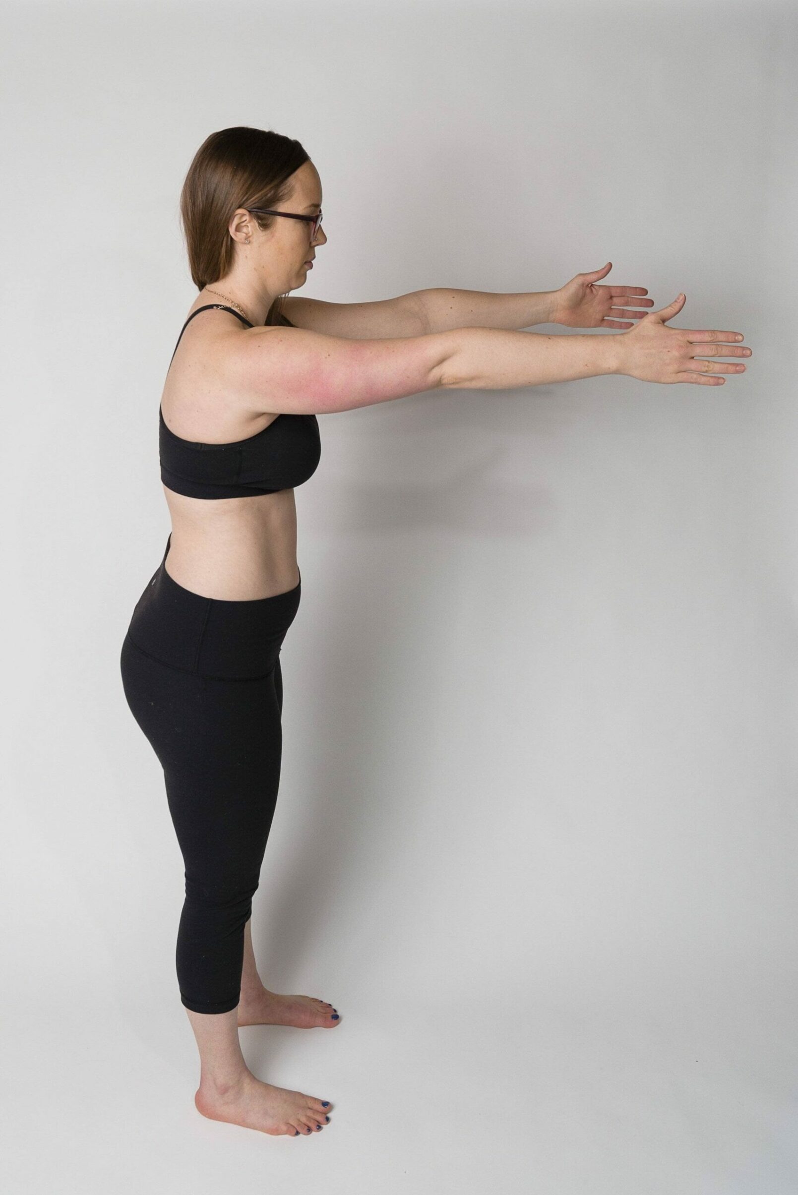 a woman standing facing to the side, with her arms straight out in front of her at shoulder height. She's demonstrating the "ribs over hips" body position for exercising after c-section.